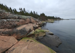 Punertavanharmaat, levien ja jäkälien koristamat silokalliot laskeutuvat tyynipintaiseen Itämereen harmaan taivaan alla.