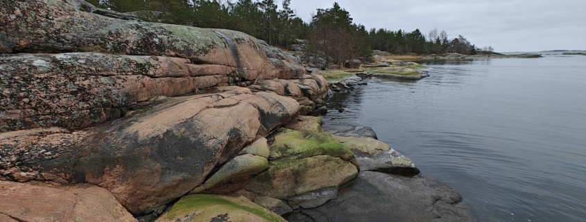 Punertavanharmaat, levien ja jäkälien koristamat silokalliot laskeutuvat tyynipintaiseen Itämereen harmaan taivaan alla.